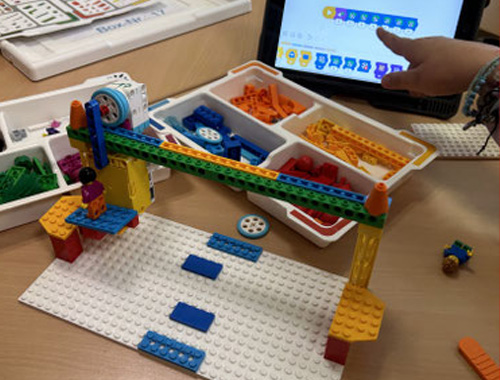 Lego-Modell auf einer weißen Grundplatte mit Figuren und Bausteinen, daneben ein Tablet mit Programmieroberfläche.