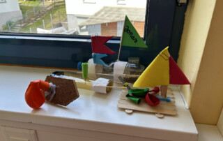 Mehrere bunte selbstgebastelte Modellboote mit Segeln aus Papier und Holz stehen auf einer weißen Fensterbank vor einem Fenster.