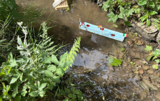 Kind steht am Bachufer und setzt ein Modellboot mit Segel ins Wasser.