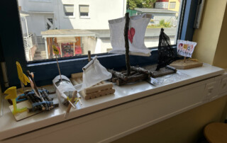 Selbstgebaute kleine Modellboote aus Holz, Korken und Papiersegeln stehen auf einer Fensterbank vor einem Fenster.