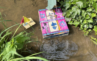 Selbstgebautes Papp-Boot mit Mast und Segel steht auf einem Tisch, daneben ein Modellboot aus Kork.