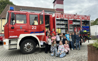 Gruppe von Kindern steht vor einem Feuerwehrwagen auf einem gepflasterten Platz.