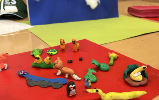 Mehrere bunte Knetfiguren liegen auf einer roten Unterlage vor einer Bastelkulisse.