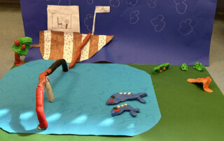 Bastelszene mit einer blauen Wasserfläche aus Stoff, Knetfiguren und einer Kulisse aus Karton.