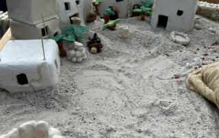 Modelllandschaft aus Sand mit weißen Figuren und kleinen Modellhäusern im Hintergrund.