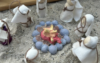 Miniaturszene in Sand mit kleinen weißen Figuren, die um ein bunt gestaltetes Lagerfeuer sitzen.