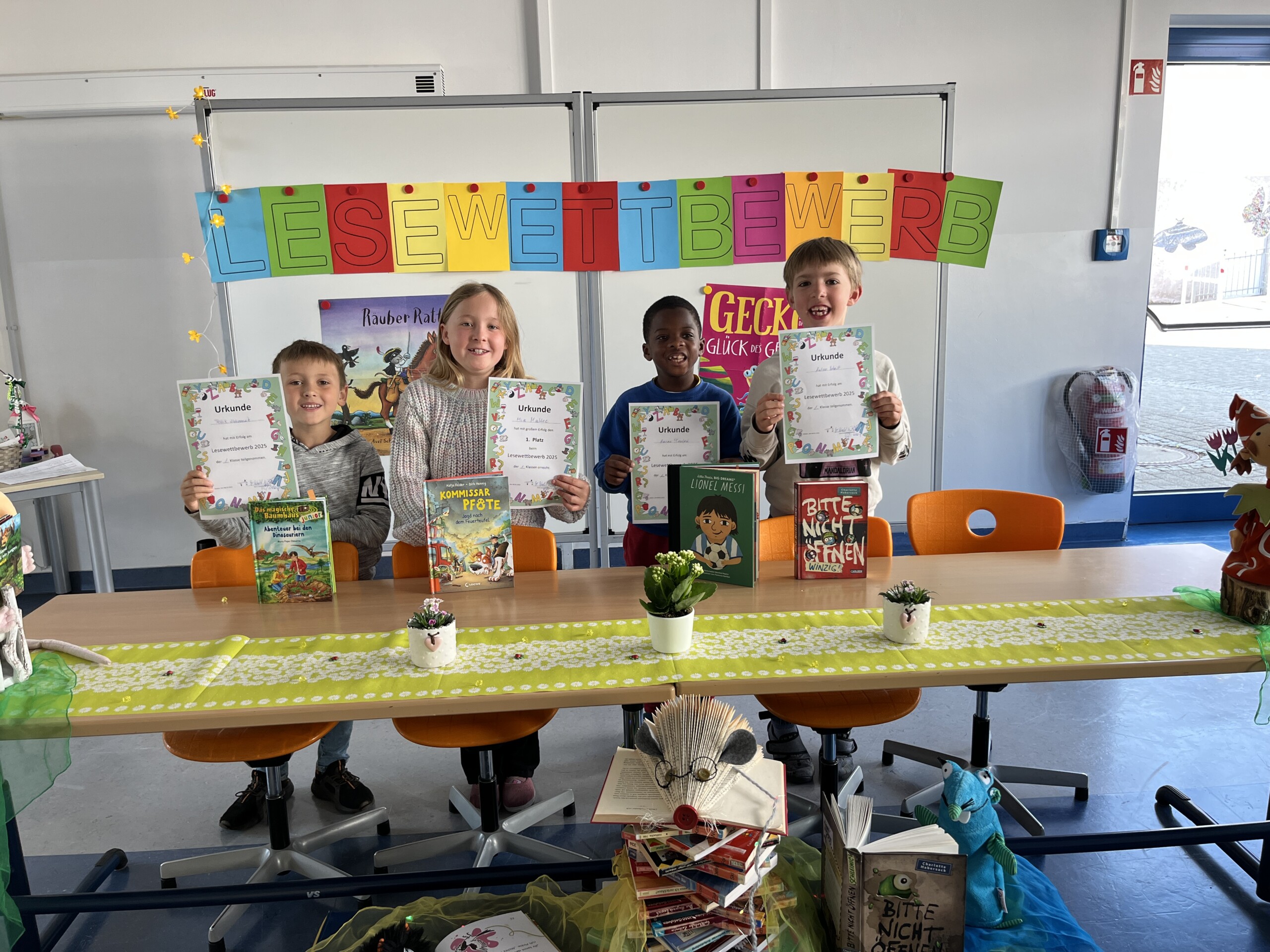 Kinder stehen im Klassenraum vor einer bunten Wanddekoration und halten ihre Arbeiten hoch.