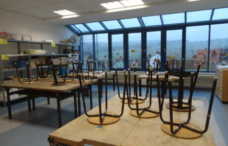 Innengebäude Klassenraum mit hochgestellten Stühlen auf Tischen und großen Fenstern an der Stirnseite.