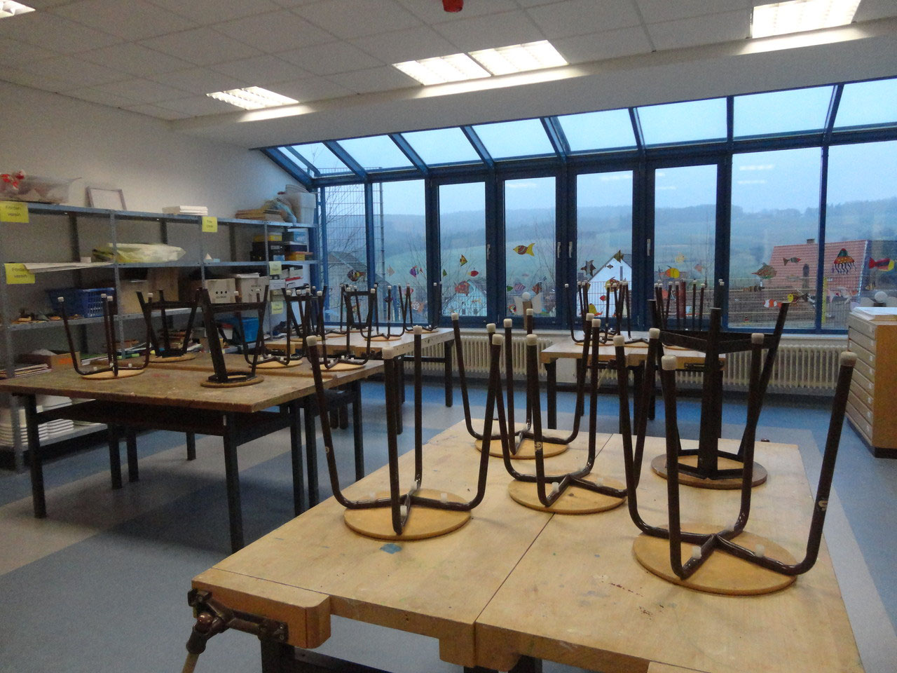 Klassenraum mit hochgestellten Stühlen auf Tischen und großen Fenstern an der Stirnseite.