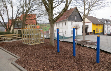 Pausenhof Spielplatz mit Klettergerüst und blau-gelben Pfosten auf Rindenmulch.