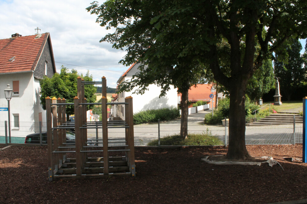 Spielplatzbereich mit Klettergerüst neben schattigen Bäumen.