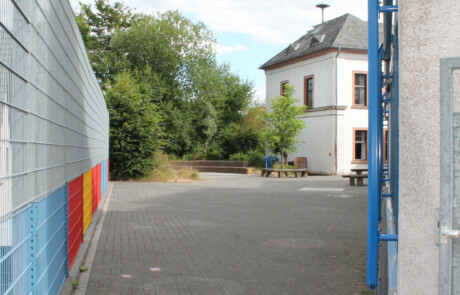 Pausenhof Gepflasterter Weg zwischen Schulgebäuden mit orange-blauer Wand und Blick auf den Schulhof.