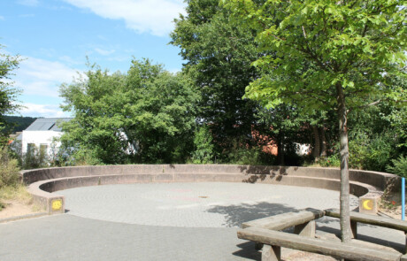 Pausenhof Schulhofbereich mit Bäumen und einer Sitzinsel aus Holzstämmen.