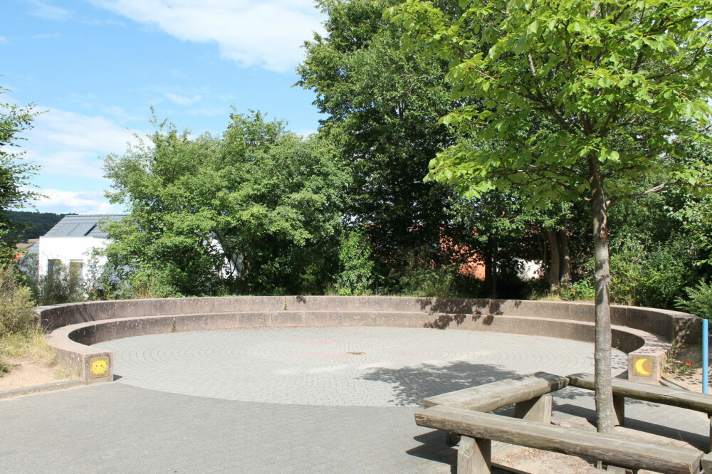 Schulhofbereich mit Bäumen und einer Sitzinsel aus Holzstämmen.