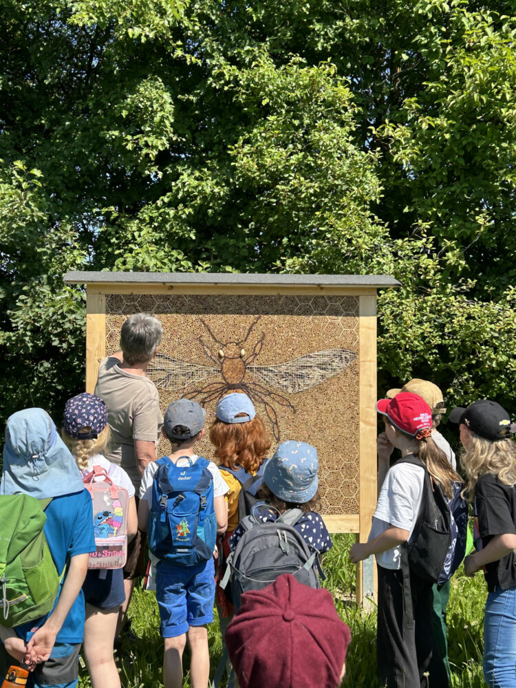 Projekt Insektenhotel Kinder stehen im Freien vor einer großen Holztafel mit einer gezeichneten Biene.