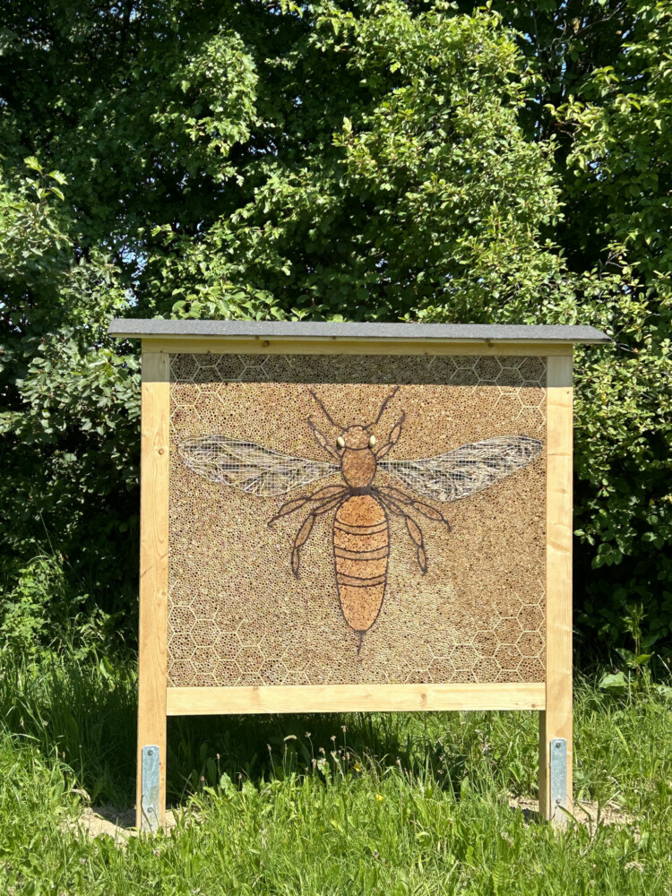 Projekt Insektenhotel Holztafeln mit einer großen gezeichneten Biene stehen auf einer Wiese vor Bäumen.