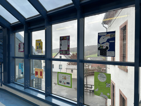 Fenster eines überdachten Verbindungsgangs mit verschiedenen farbigen Plakaten, die nach außen sichtbar angebracht sind.