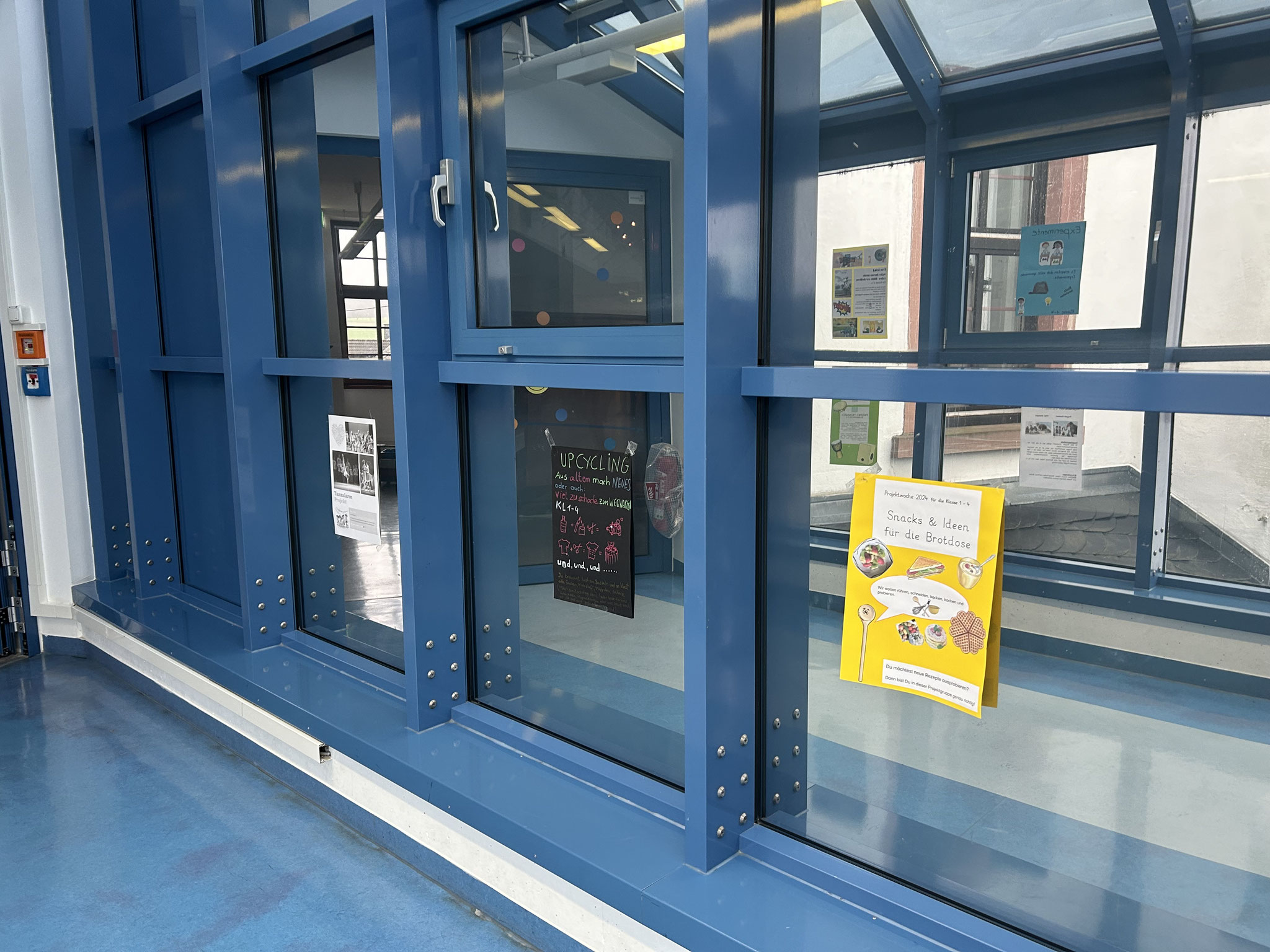 Projektwoche und Schnuppertag_3 Fensterfront eines Schulgebäudes mit mehreren bunten Plakaten, die von innen an den Scheiben befestigt sind.