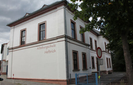 Schulgebäude_01 Seitliche Außenansicht der Alteburgschule Heftrich mit Bäumen und Schulhof.