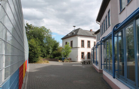 Schulgebäude_03 Außenansicht eines Schulgebäudes mit Bäumen und Metallzaun.