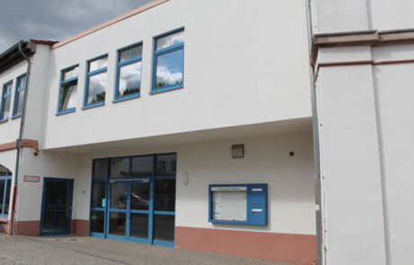 Schulgebäude_06 Schuleingang mit blauem Türbereich und überdachter Eingangszone.