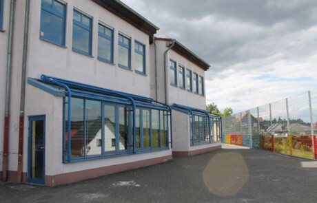 Schulgebäude_08 Außenansicht eines Schulgebäudes mit Bäumen und Metallzaun.
