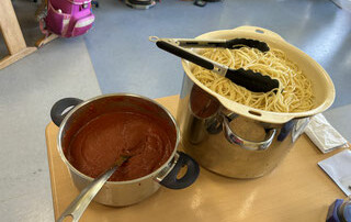 Topf mit Tomatensoße und großer Topf mit gekochten Spaghetti stehen auf einem Tisch im Klassenraum.