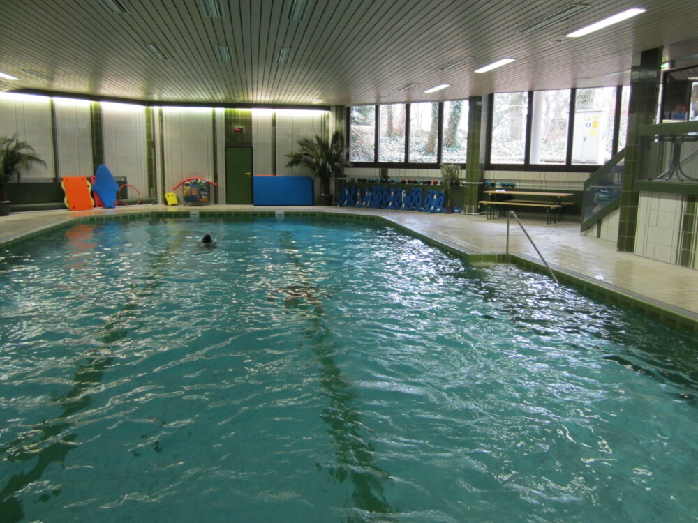 Leeres Schwimmbecken in einer Halle mit breiten Fenstern und hellen Wänden.