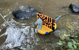 Kleine Papier- oder Holzfigur schwimmt im Bach zwischen Steinen.