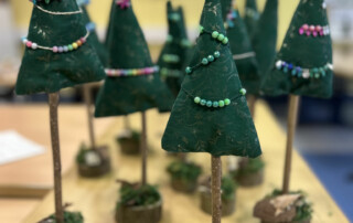 Mehrere kleine gebastelte Tannenbäume aus grünem Filz stehen auf Holzfüßen, dekoriert mit bunten Perlenketten.