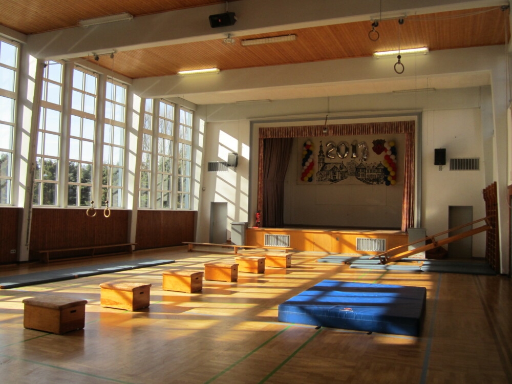 Innenraum einer Turnhalle mit Holzboden, Sportmatten und großen Fenstern mit Tageslicht.