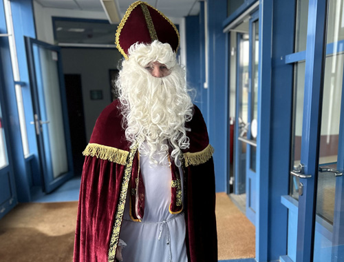 Eine Person in einem traditionellen Nikolaus- oder Bischofskostüm mit rotem Mantel, goldenen Verzierungen, weißem Bart und Mitra steht in einem Schulflur.