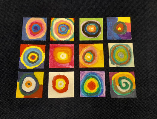 die-malerei-ag-stellt-sich-vor3 Zwölf quadratische Kunstwerke mit bunten konzentrischen Kreisen liegen auf dunklem Untergrund.