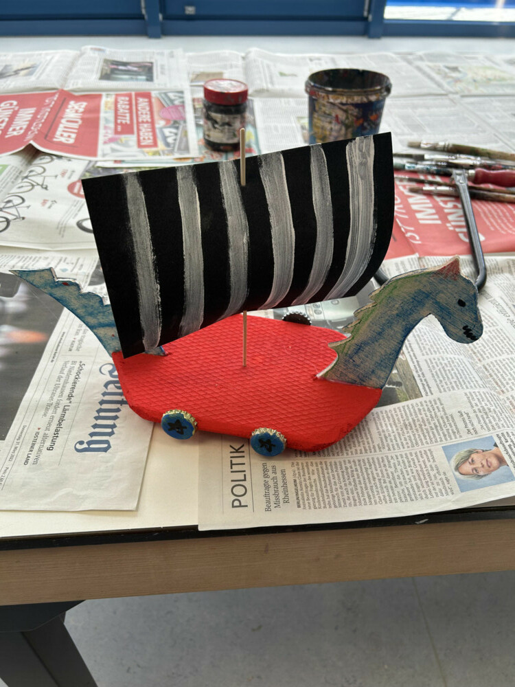 Nahaufnahme eines gebastelten roten Wikingerschiffs mit schwarz-weißem Segel und Drachenkopf-Verzierung.