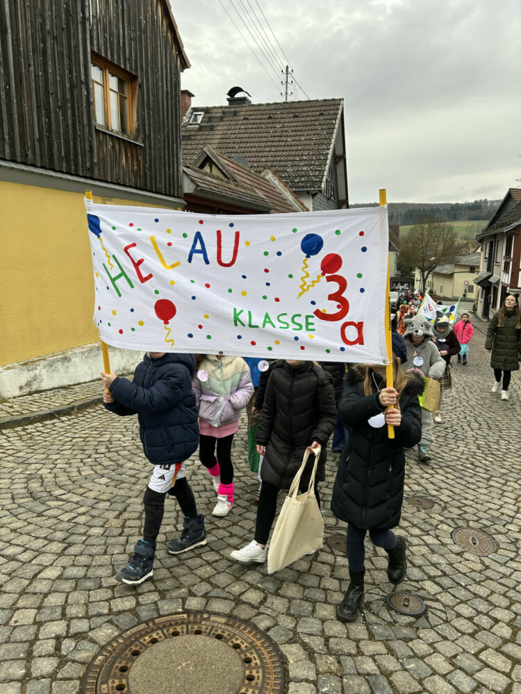 fasching-an-der-alteburgschule-heftrich1 Kinder gehen einen gepflasterten Weg entlang und halten ein großes Banner mit der Aufschrift „Helau – Klasse 3a“.