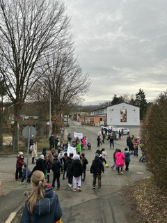 Kinder und Erwachsene gehen in einer Gruppe eine Dorfstraße entlang und tragen Plakate.