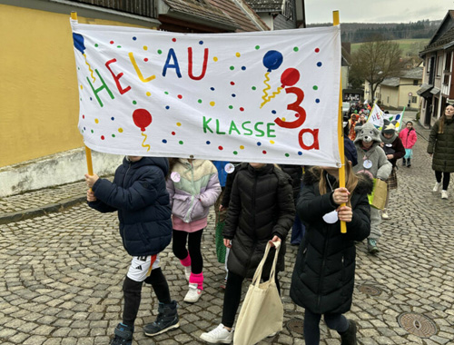 Kinder gehen einen gepflasterten Weg entlang und halten ein großes Banner mit der Aufschrift „Helau – Klasse 3a“.