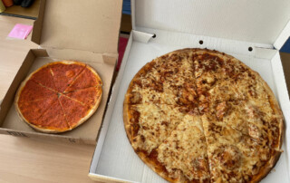 Zwei Pizzen in offenen Kartons liegen auf einem Tisch, eine große Käsepizza und eine kleinere mit Tomatenbelag.