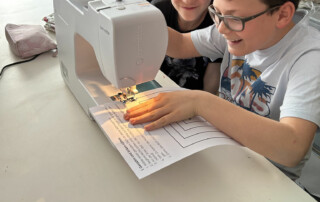 Zwei Kinder sitzen an einer Nähmaschine und arbeiten gemeinsam an einem Stoffprojekt.