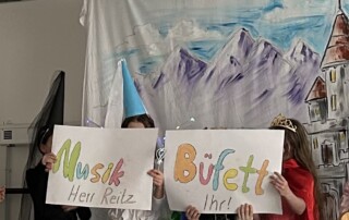 Kinder stehen vor einer Wand mit vielen aufgehängten Papierkostümen oder Bastelarbeiten.