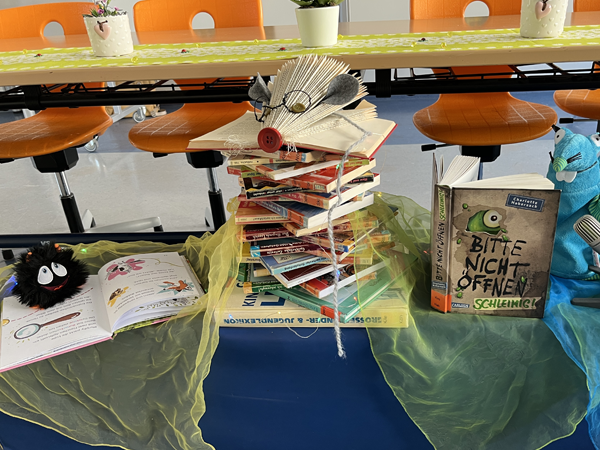 schulbücherei Dekorative Bücherpräsentation mit einem hohen Stapel Kinderbücher, einem gefalteten Buch in Schmetterlingsform, einem Plüschmonster und weiteren geöffneten Büchern auf bunten Tüchern in einem Raum mit orangefarbenen Stühlen.