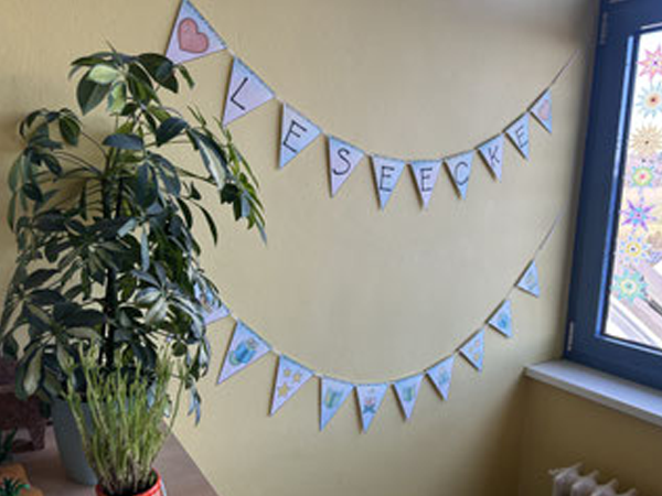 Ecke eines Klassenraums mit zwei dekorativen Wimpelketten, die das Wort „Leseecke“ bilden, und einer großen Zimmerpflanze neben einem Fenster.