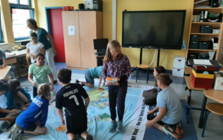 Kinder stehen und sitzen um eine große Lernplane mit Waldmotiven und bearbeiten Aufgaben darauf.