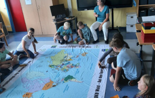 Kinder arbeiten gemeinsam auf einer großen Lernplane mit Natur- und Waldmotiven im Klassenraum.