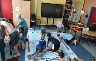 Kinder sitzen in einem Klassenraum auf einer großen Lernplane, die ein Wald- oder Naturthema zeigt.